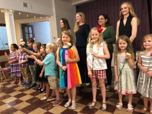Children holding flowers on stage, with 4 adult women behind them