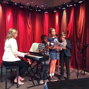 Woman playing keyboard while 3 children sing into microphone