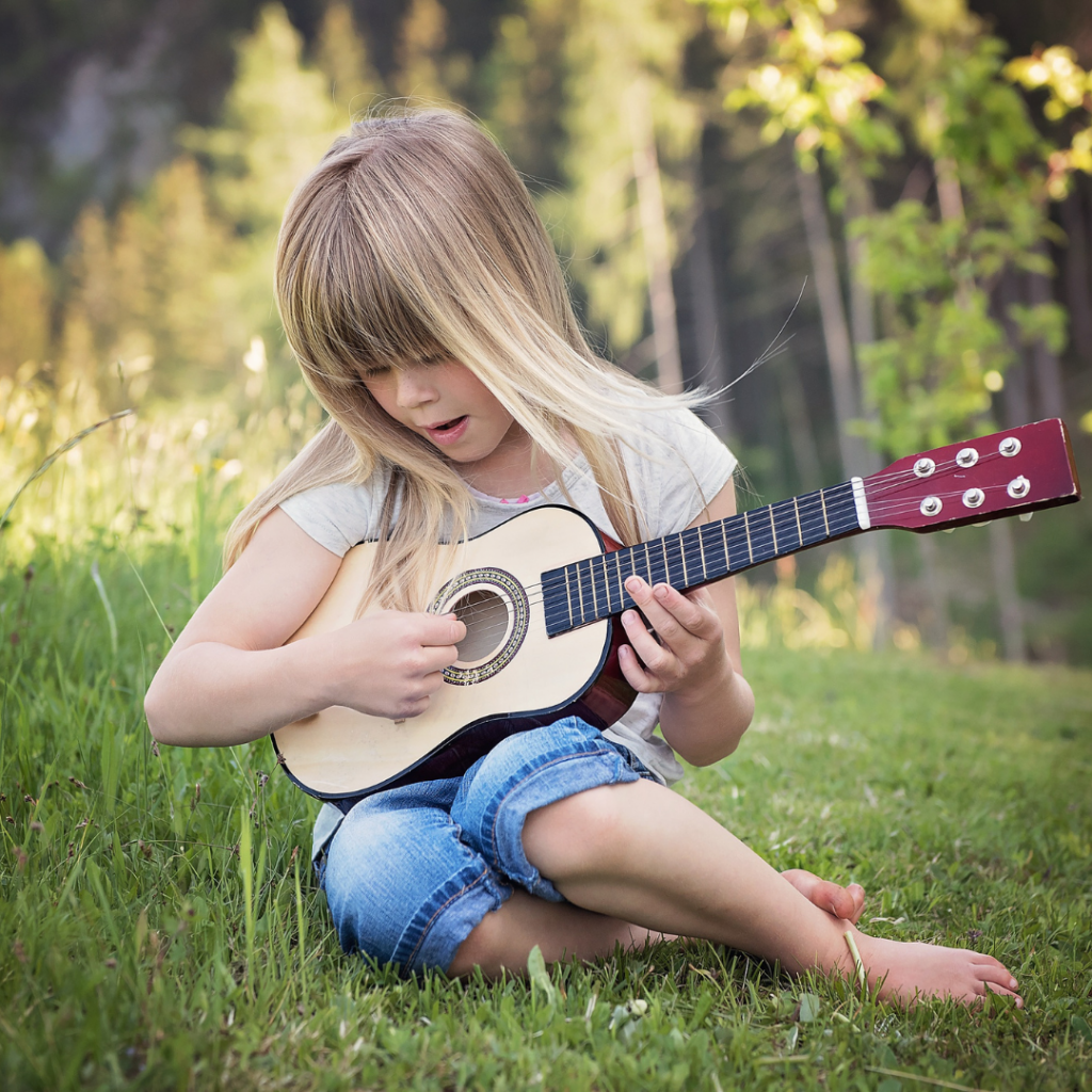 Blond girl in jean shorts sitting on the ground outside playing a ukulele