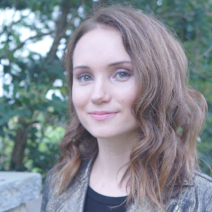 Pretty woman with wavy brown hair smiling, trees in background
