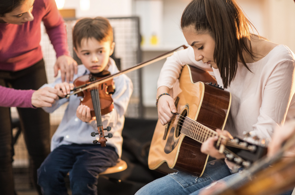 Two people being instructed while playing instruments. A boy with a violin and a young woman with an acoustic guitar.