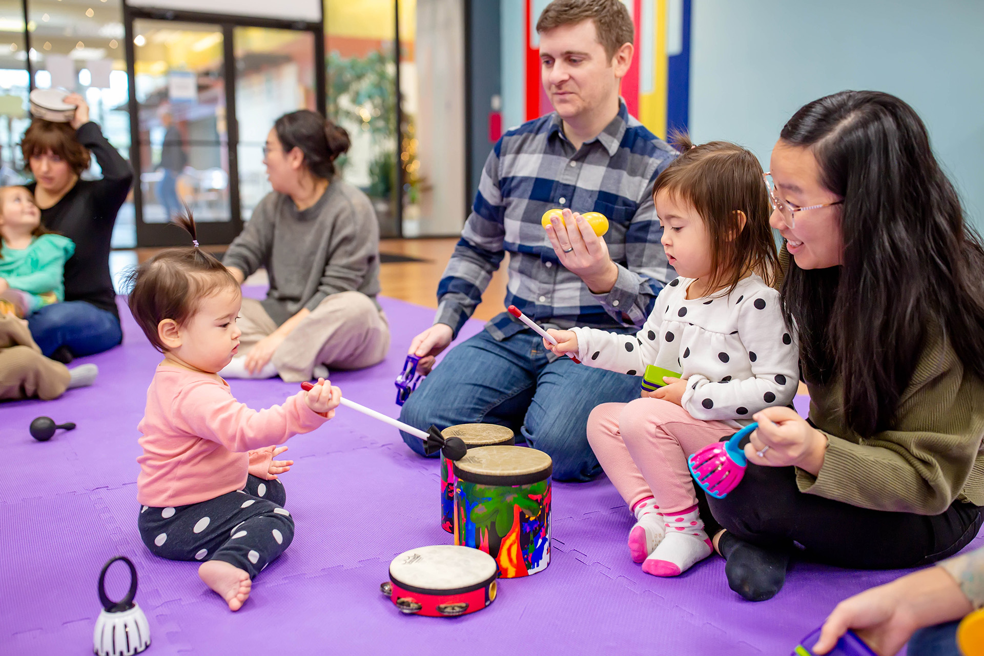 A mom and dad sit on a purple carpet with their two children. The children are playing small drums with wooden mallets, everyone is smiling.
