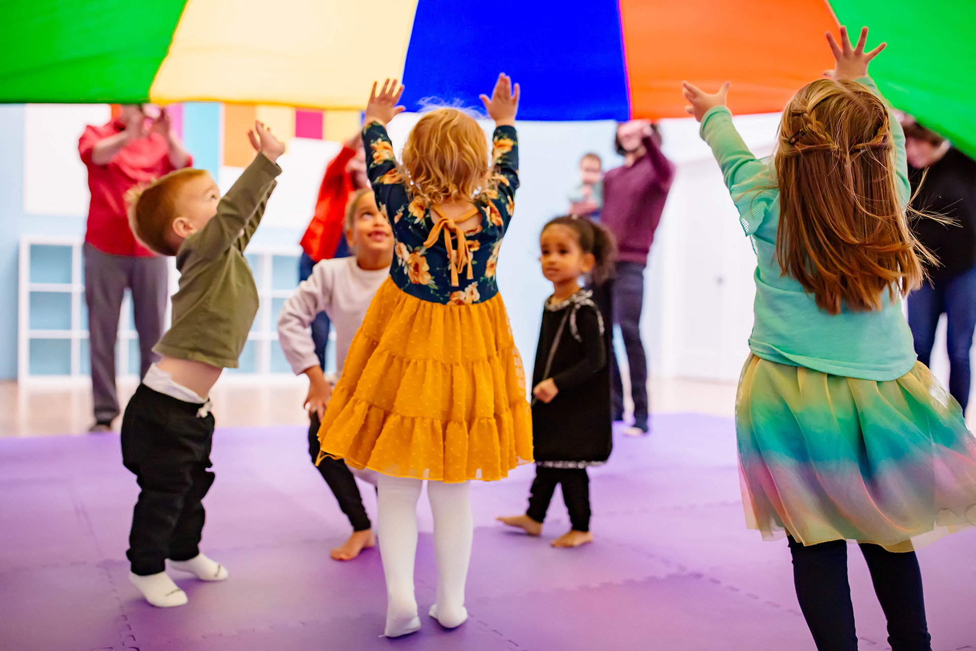 A group of toddlers standing on a purple rug lift a multi-colored parachute high over their heads.