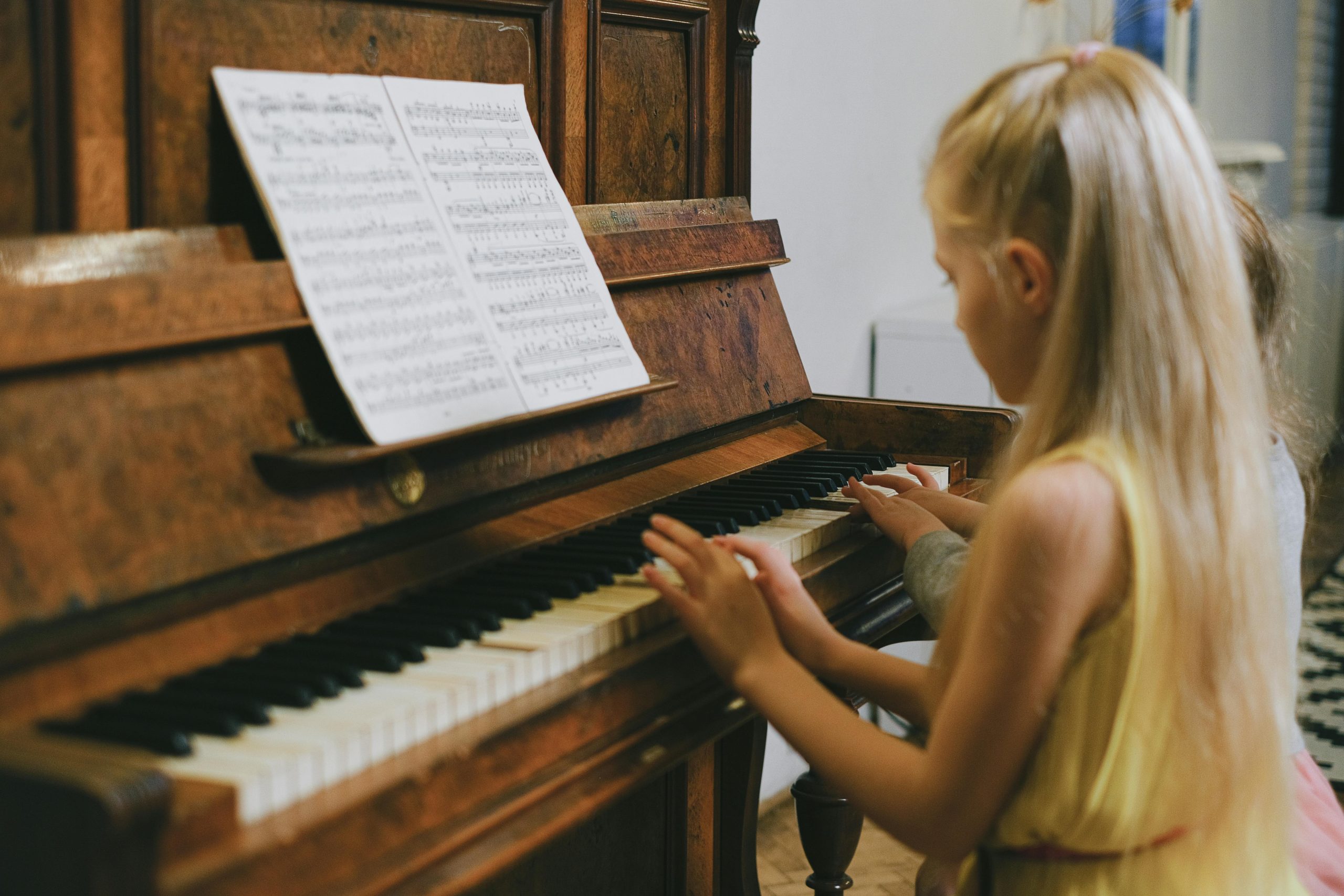 Little girl sits at a wooden upright piano, appearing to play from the sheet music propped on the stand.