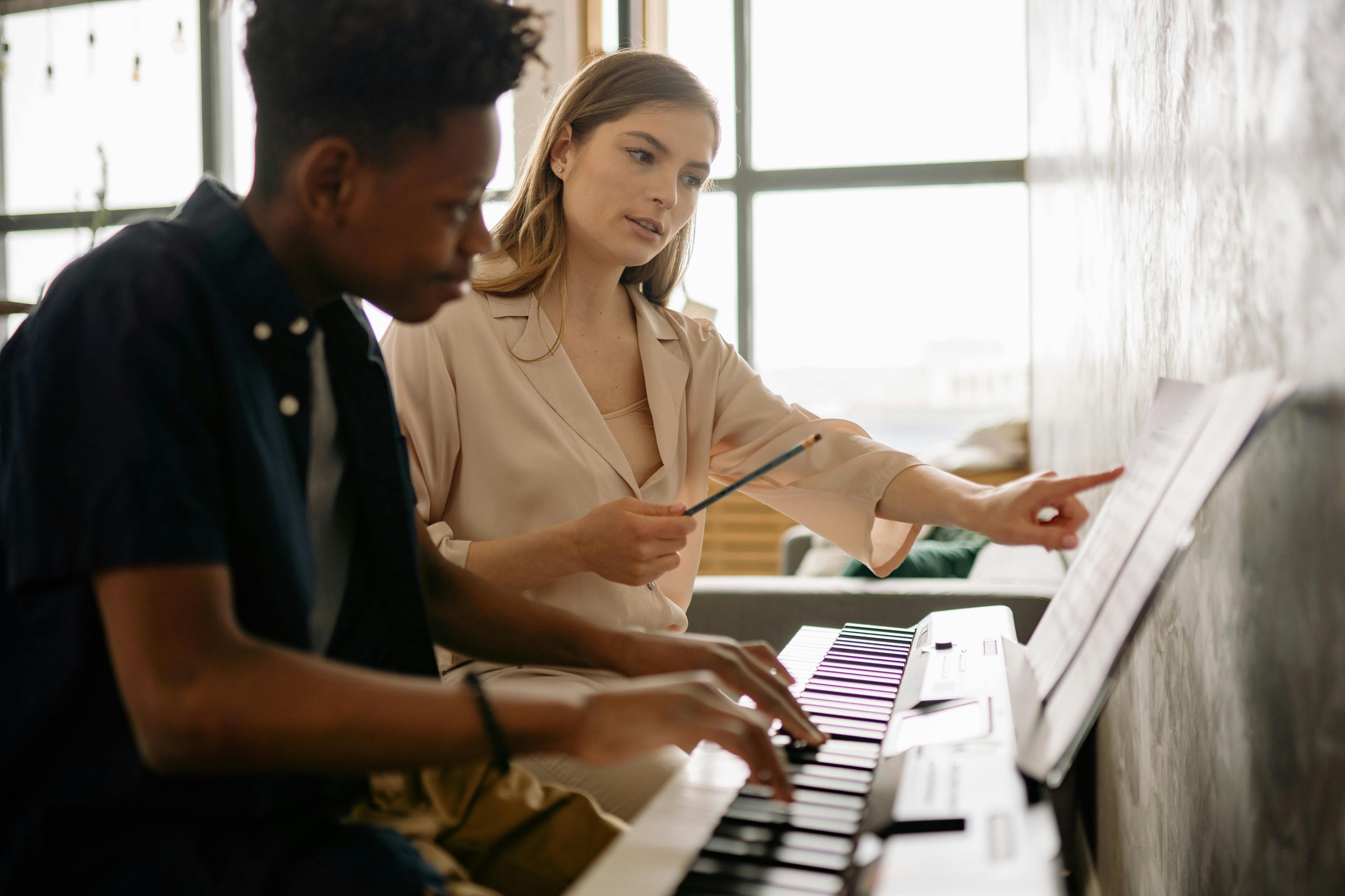 A young woman with fair skin and long brown hair sits next to her piano student. He has brown skin and short curly black hair, he looks intently at the keys.